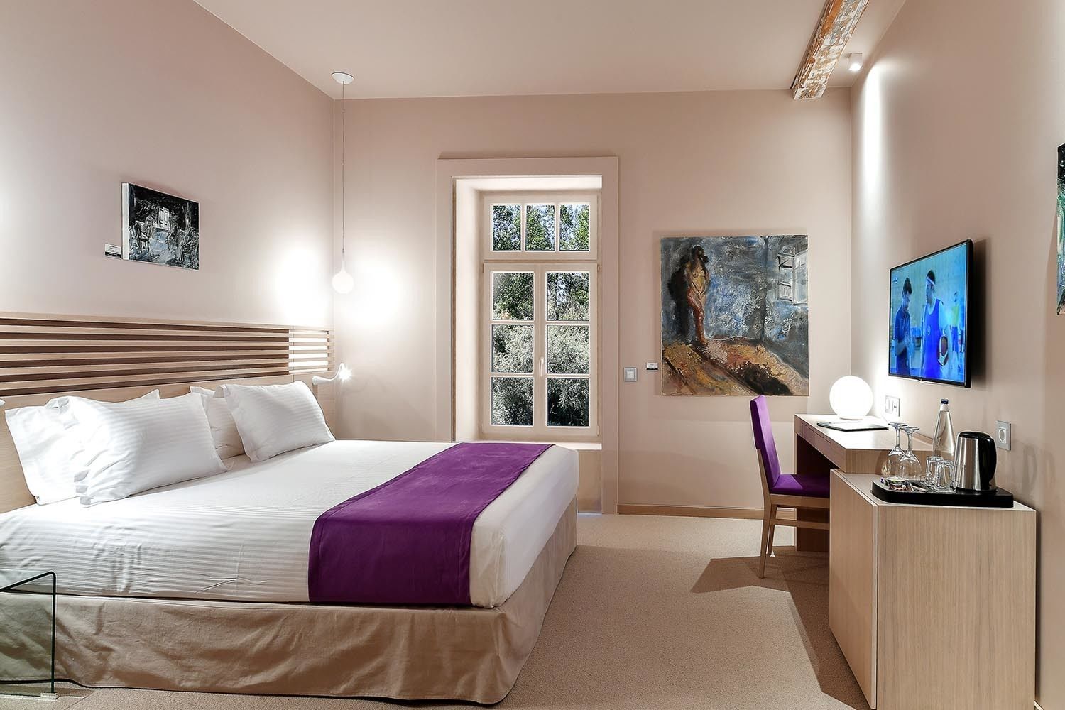 A modern hotel room with a bed, a desk, a chair, a TV, and artwork on the walls.