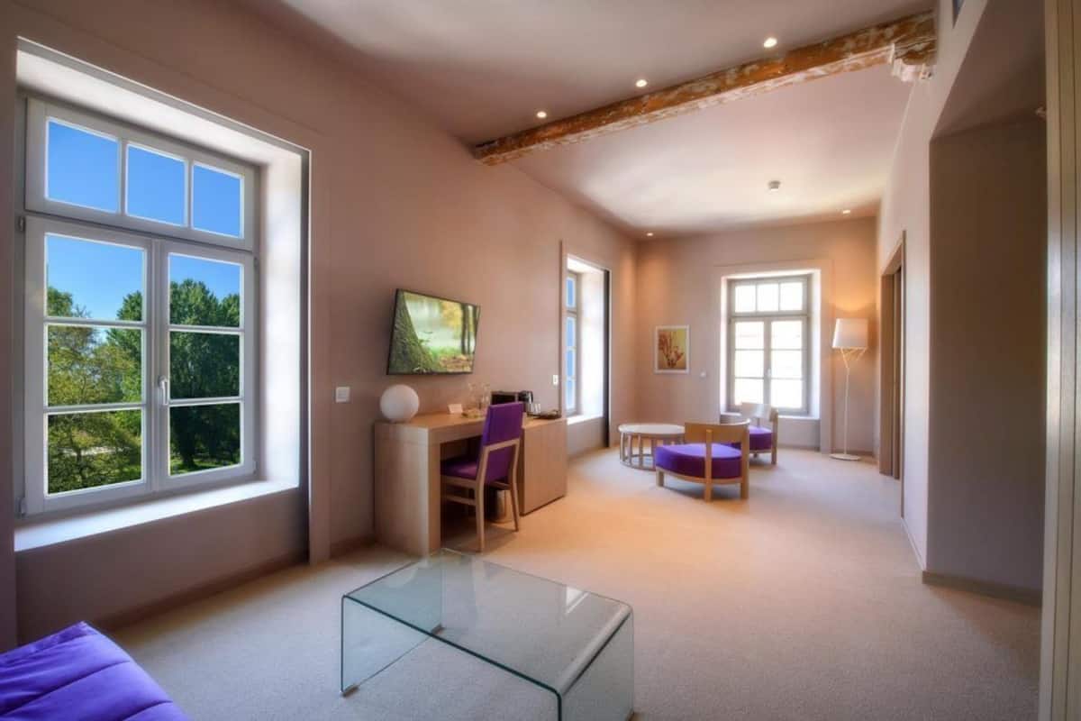 Ein geräumiges Zimmer mit großen Fenstern, einem Glastisch, einem Schreibtisch mit Stuhl und einem violetten Sofa.