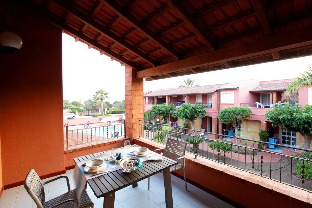 A balcony with a table set for dining, surrounded by chairs, overlooking a residential area with houses and greenery.