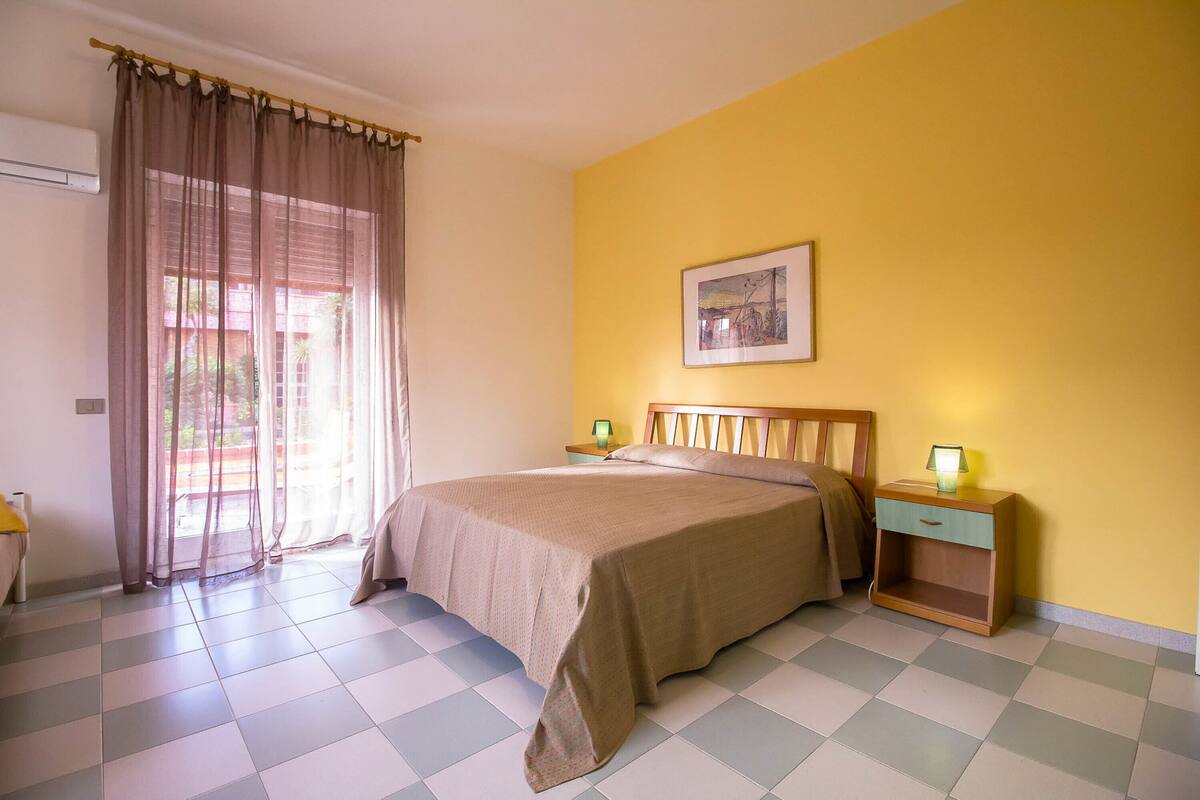 A bedroom with a bed, a nightstand, a lamp, a window with curtains, and a wall-mounted picture.