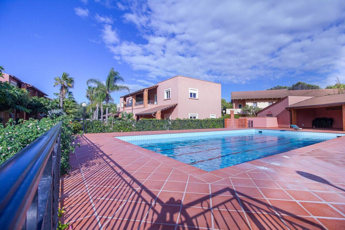 A swimming pool with a tiled deck, surrounded by buildings with balconies and a clear blue sky.