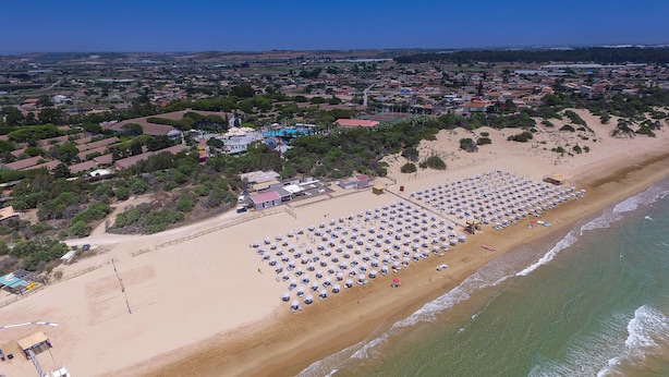An aerial view of a beach with rows of sunbeds and umbrellas, a sandy shoreline, and a backdrop of residential buildings and greenery. - Residence del Mare - Case Sicule (Ispica)