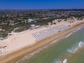 An aerial view of a beach with rows of sunbeds and umbrellas, a sandy shoreline, and a backdrop of residential buildings and greenery. - Residence del Mare - Case Sicule (Ispica)