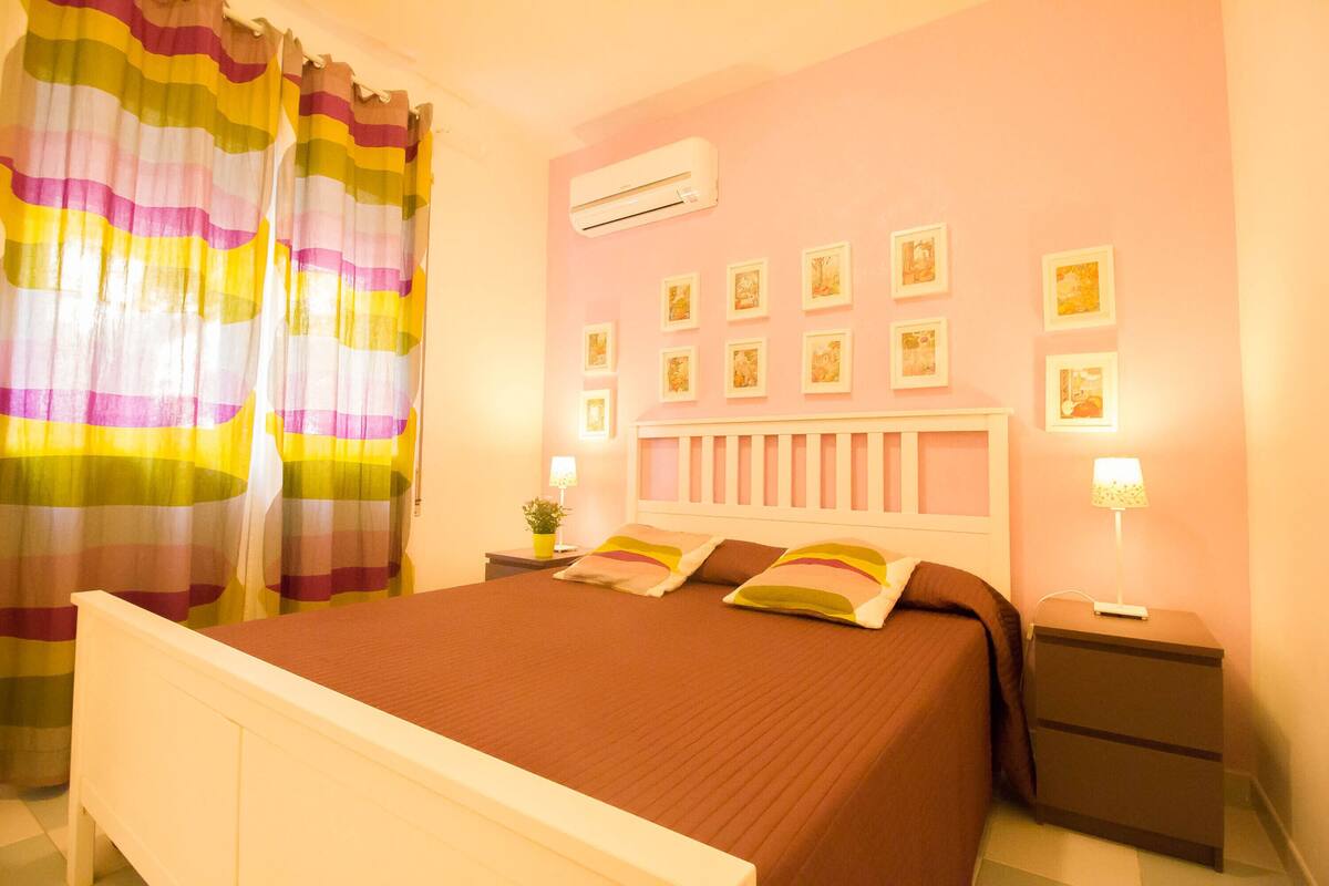 A bedroom with a bed, nightstands, a wall-mounted air conditioner, and framed pictures on the wall.