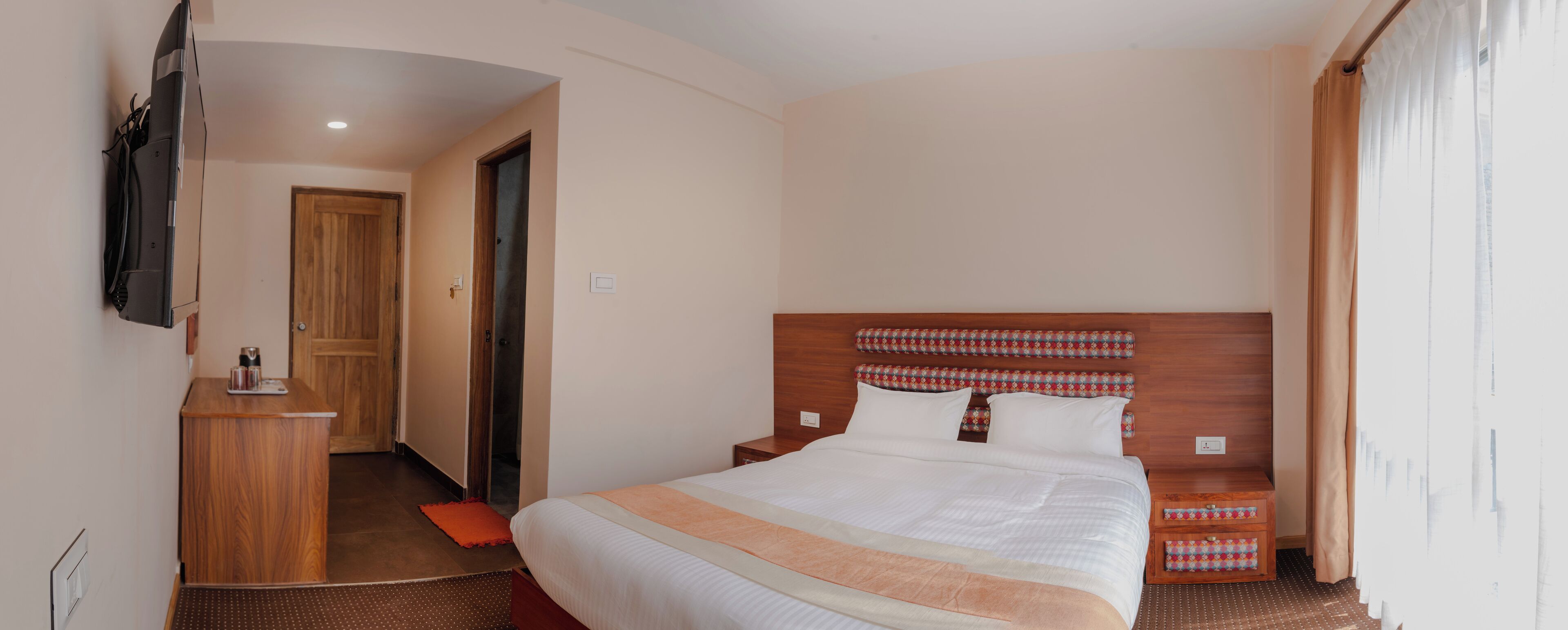 Quarto de hotel com cama grande, cabeceira de madeira, pequena escrivaninha e televisão fixada na parede.