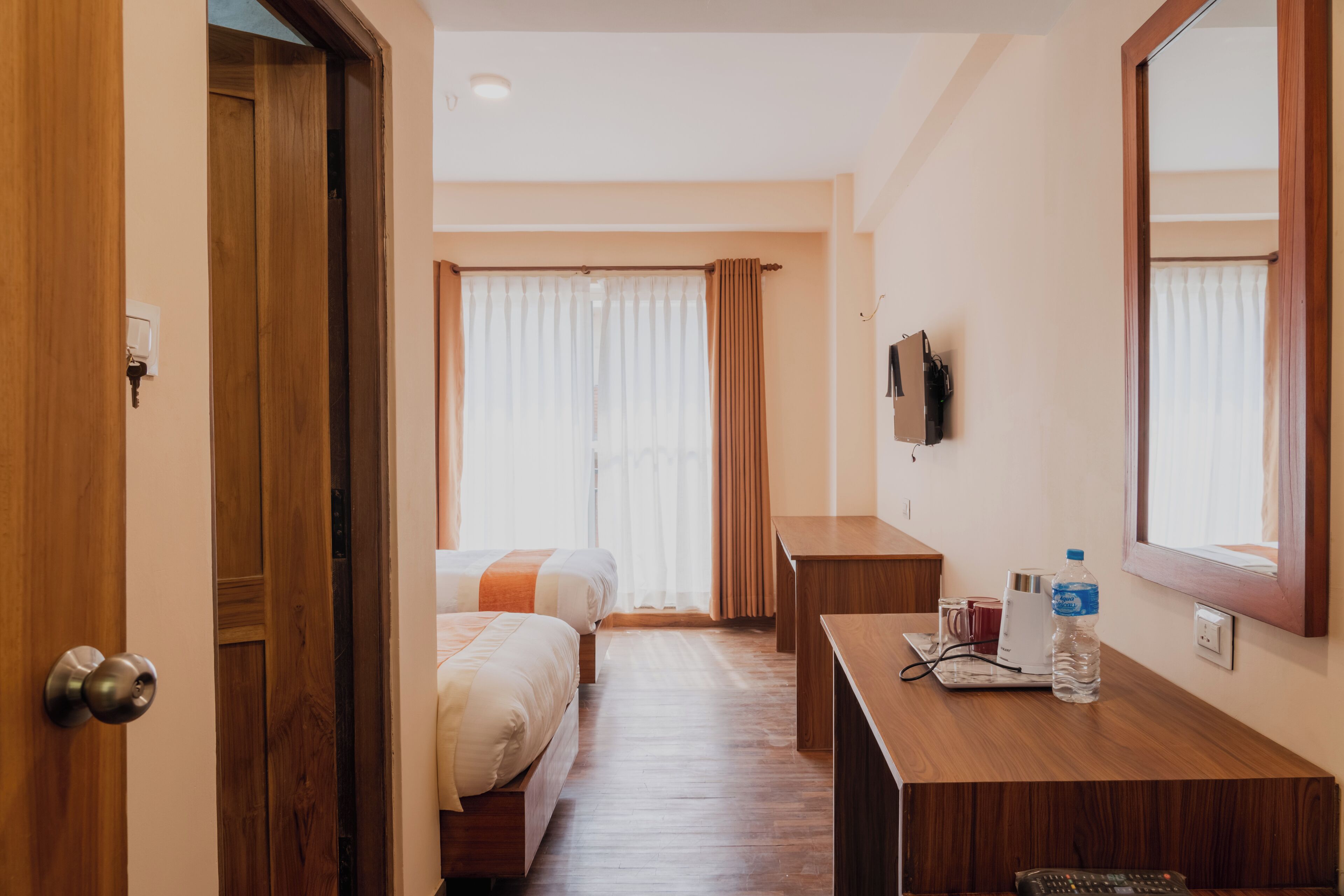 Una habitación de hotel con dos camas, un escritorio, un espejo, un televisor y una ventana con cortinas.