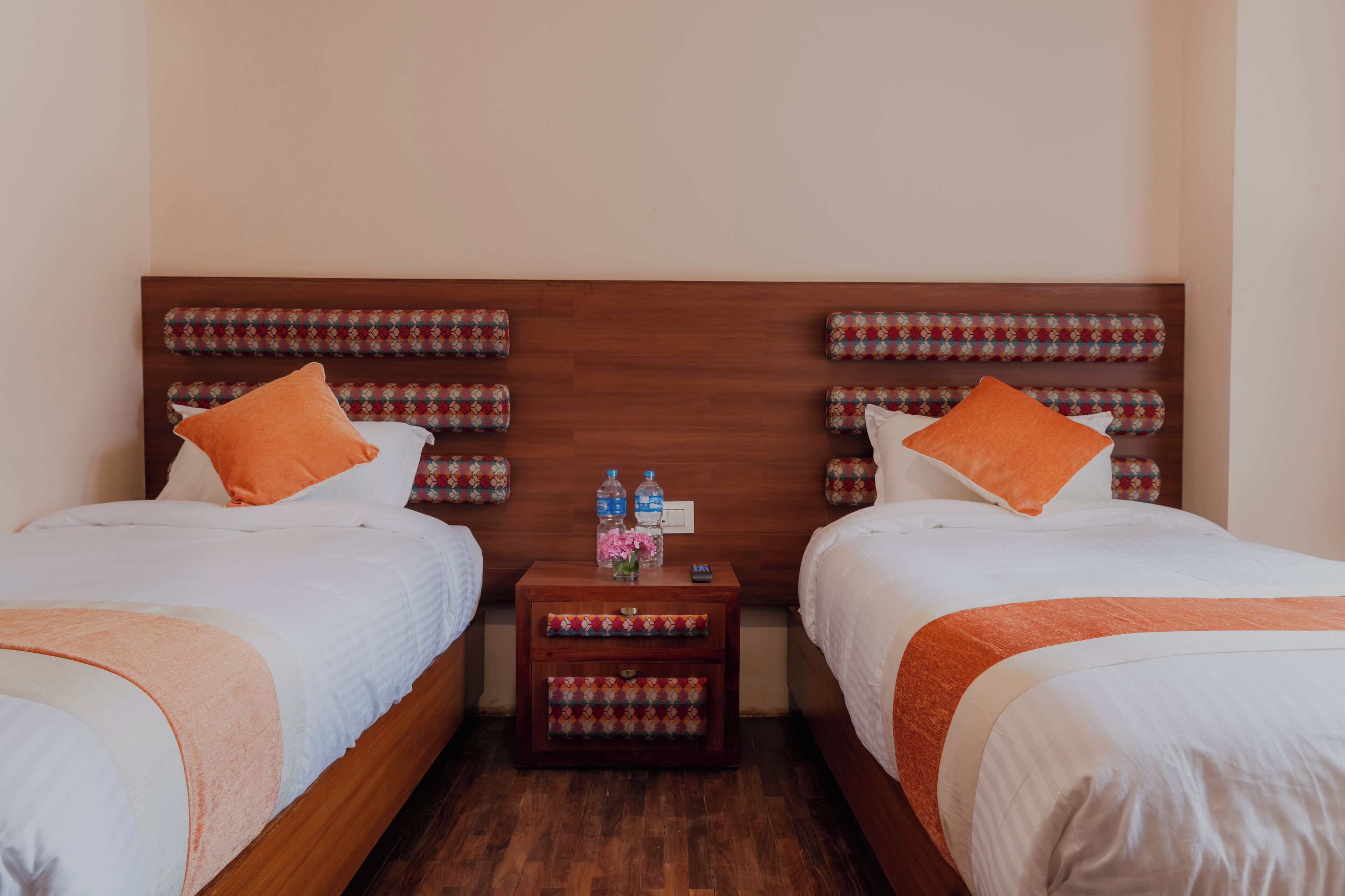 A double room with two beds, each with an orange pillow, a wooden headboard, and a small bedside table with a floral vase and water bottles.