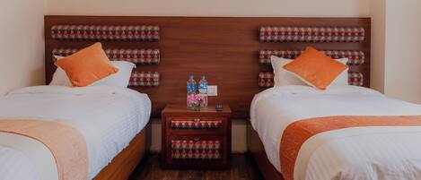 A double room with two beds, each with an orange pillow, a wooden headboard, and a small bedside table with a floral vase and water bottles.