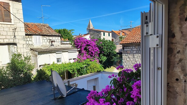 Parveke, jossa on satelliittiantenni, kivirakenteinen rakennus, jonka katto on päällystetty tiilillä, ja violetit bougainvillea-kasvit.