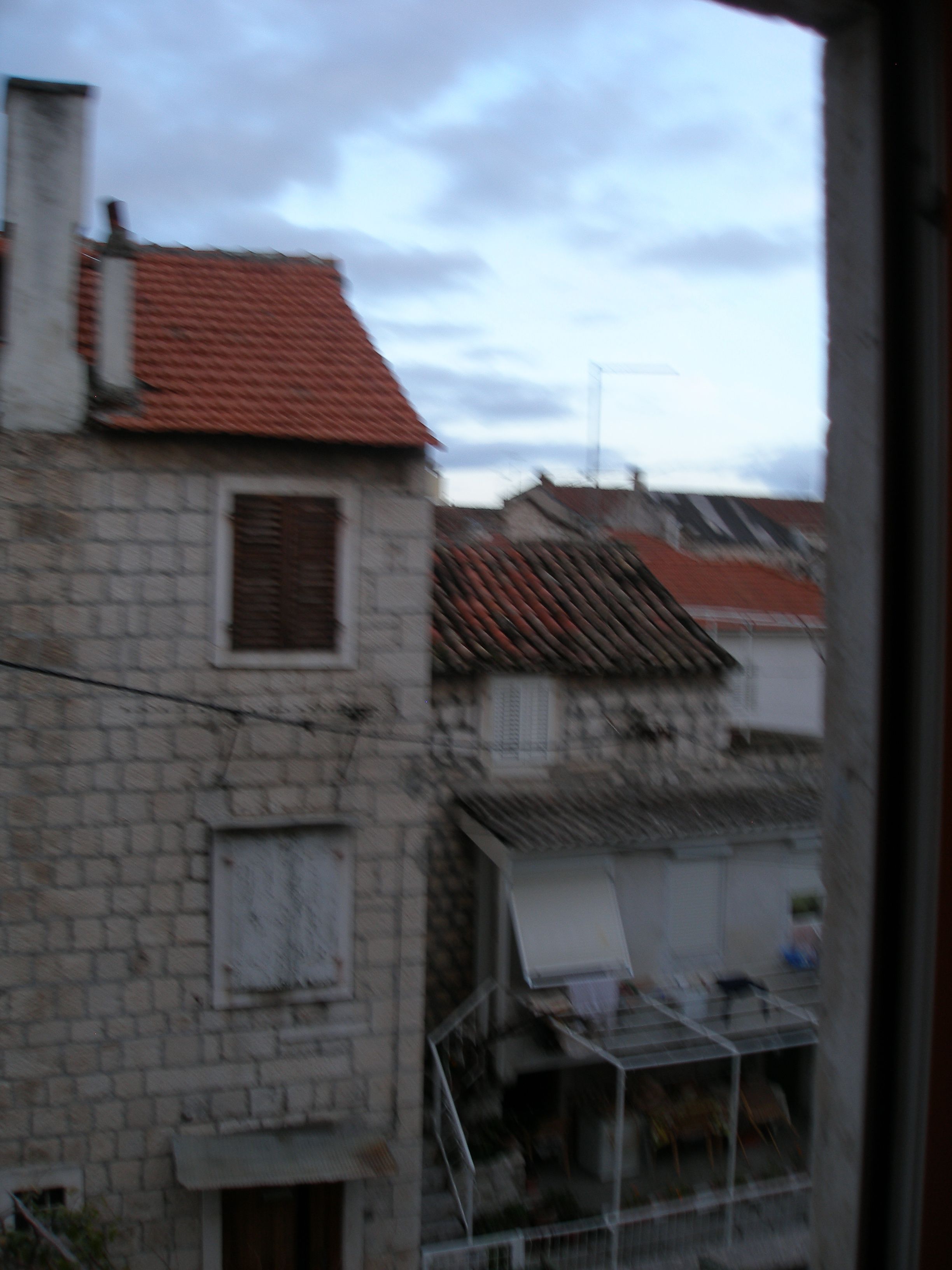 A view of a stone building with a red-tiled roof, a window with shutters, and a metal fence.