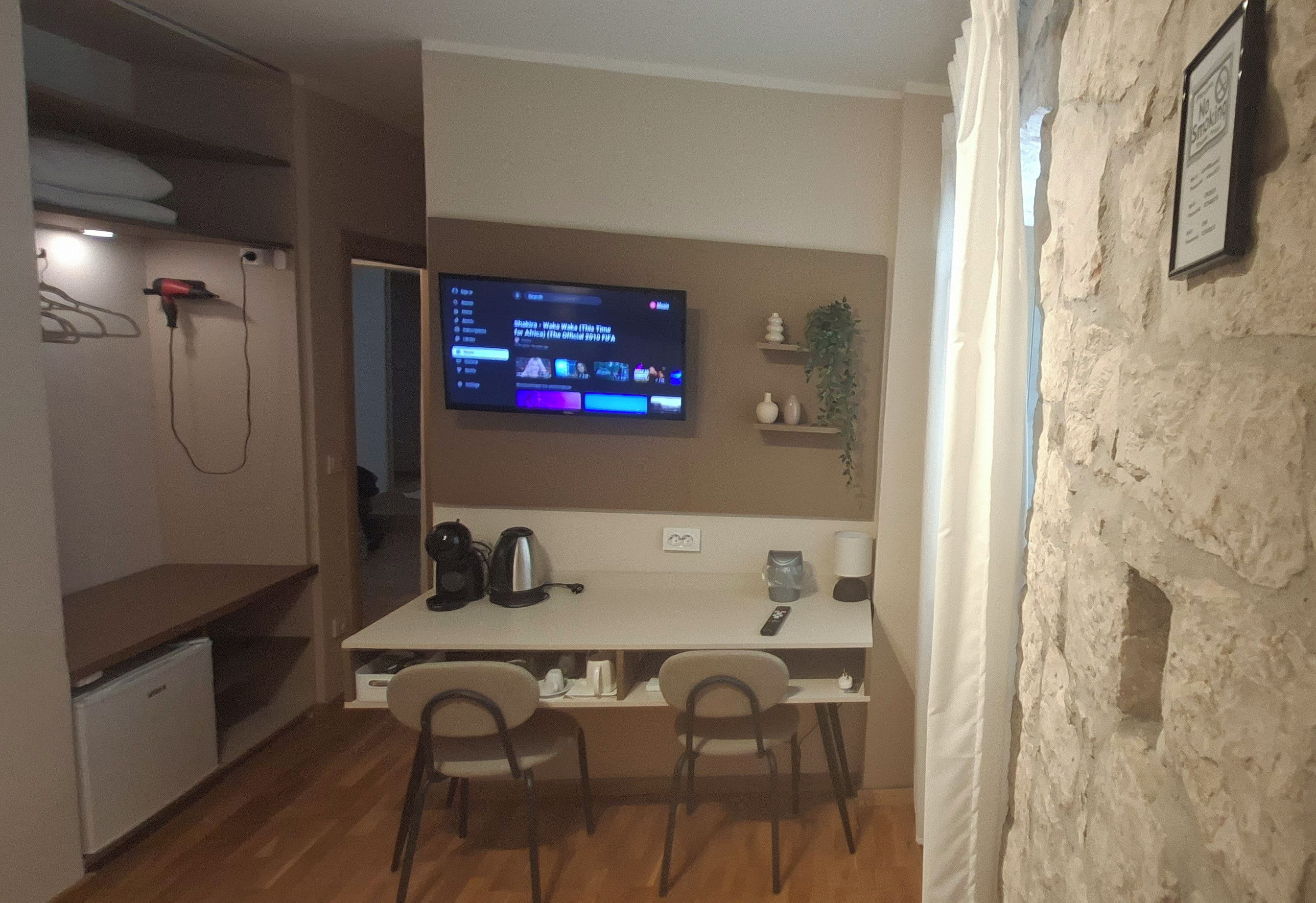 Ett kompakt boendeutrymme med ett litet kök, en matplats med två stolar, en tv som är monterad på väggen och en liten växt på en hylla.