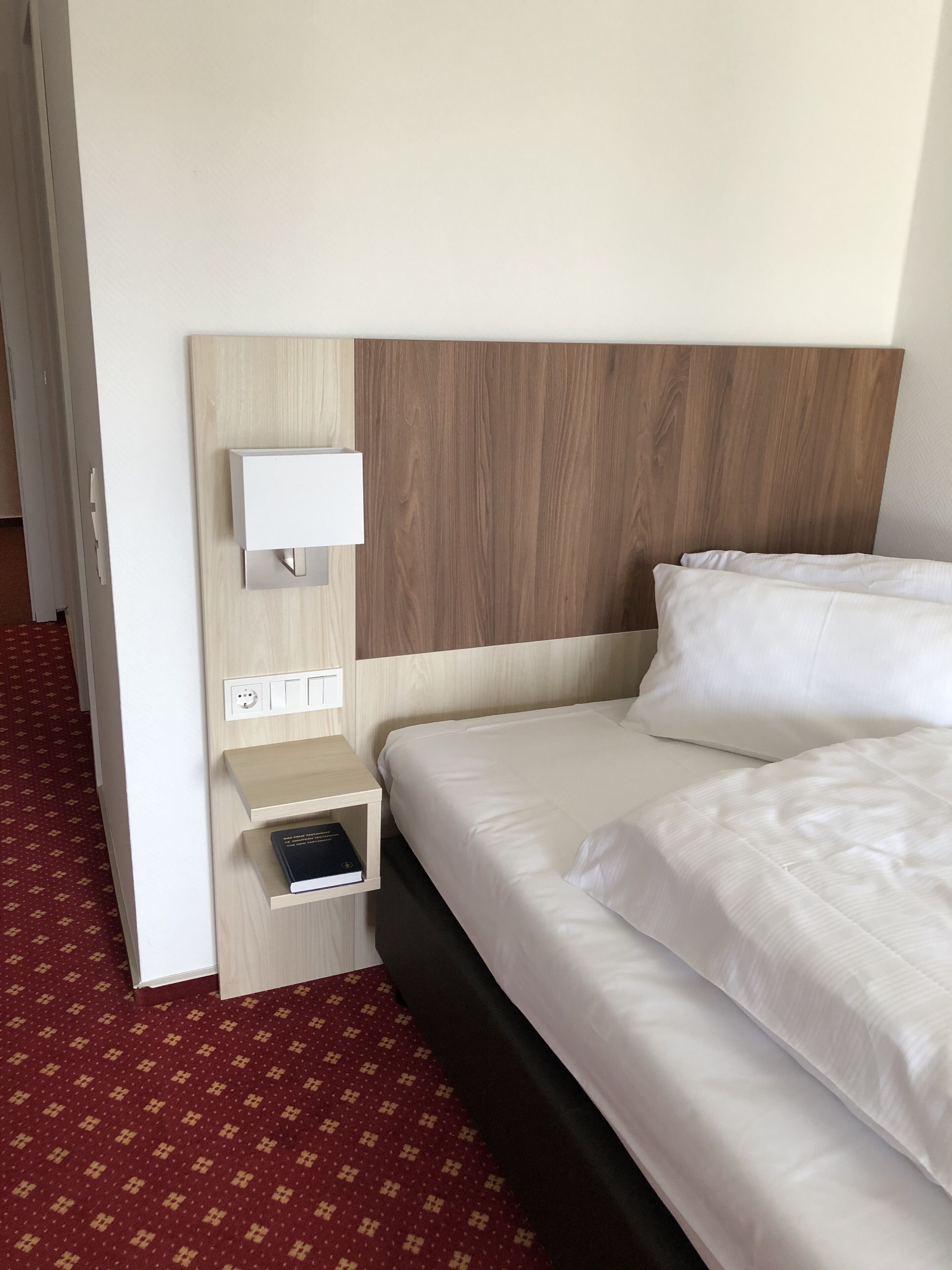 A hotel room with a wooden headboard, a bedside table with a book, and a red patterned carpet.