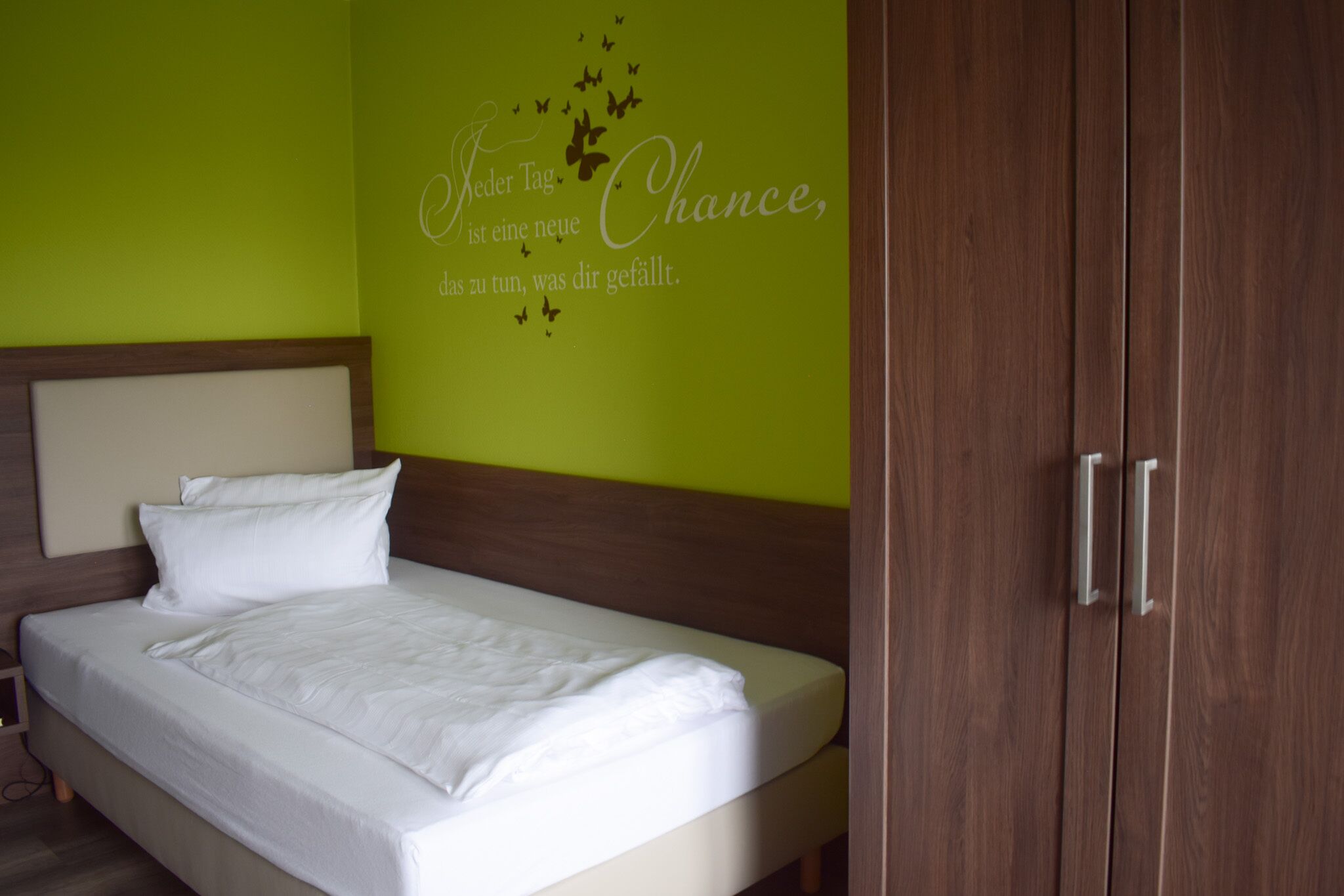 A neatly made bed with white linens in a room with a green wall and a wooden wardrobe.