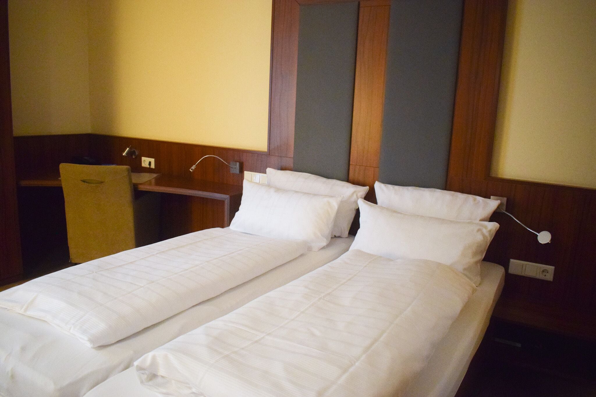 Habitación de hotel con dos camas individuales, cabecero de madera, escritorio con silla y un espejo.