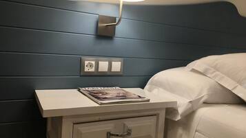 Una cama bien hecha con ropa blanca, una mesita de noche con una revista y una lámpara de pared.