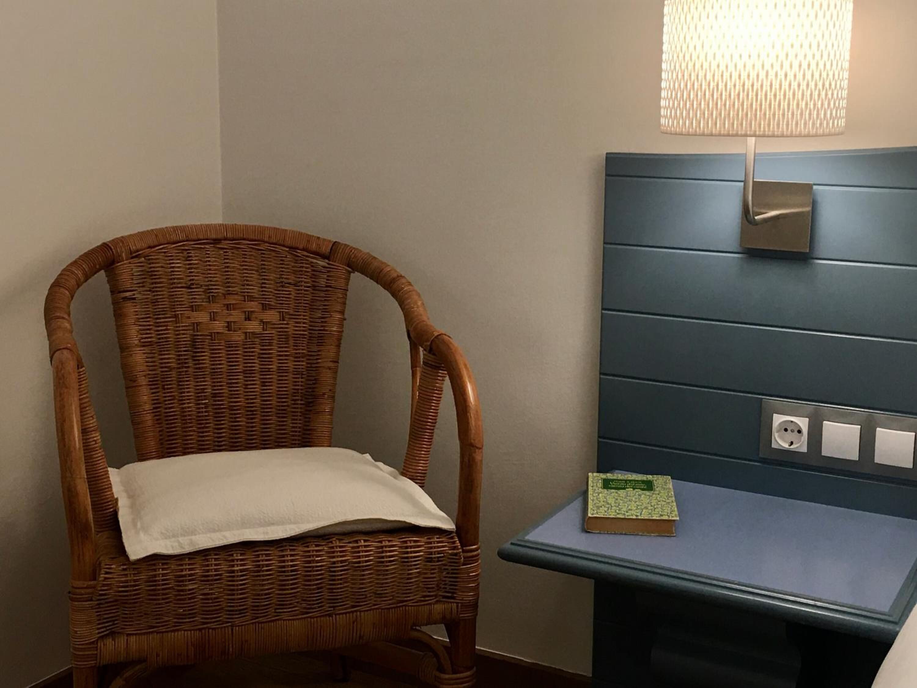 Une chaise en osier avec un coussin blanc, une petite table sur laquelle repose un livre et une lampe fixée au mur.