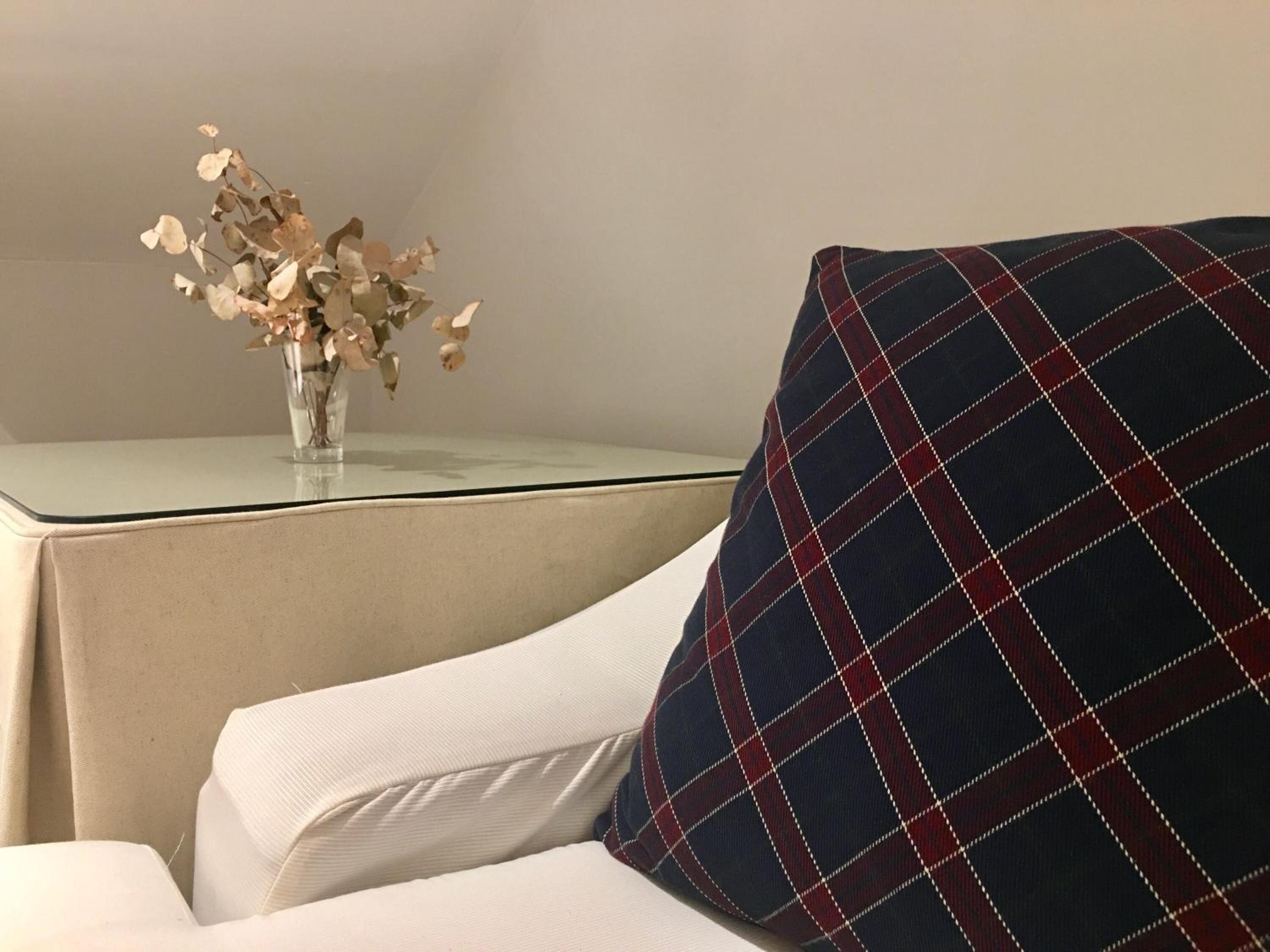 Un rincón de una habitación con un sillón blanco, una mesa de vidrio y un florero con flores secas.