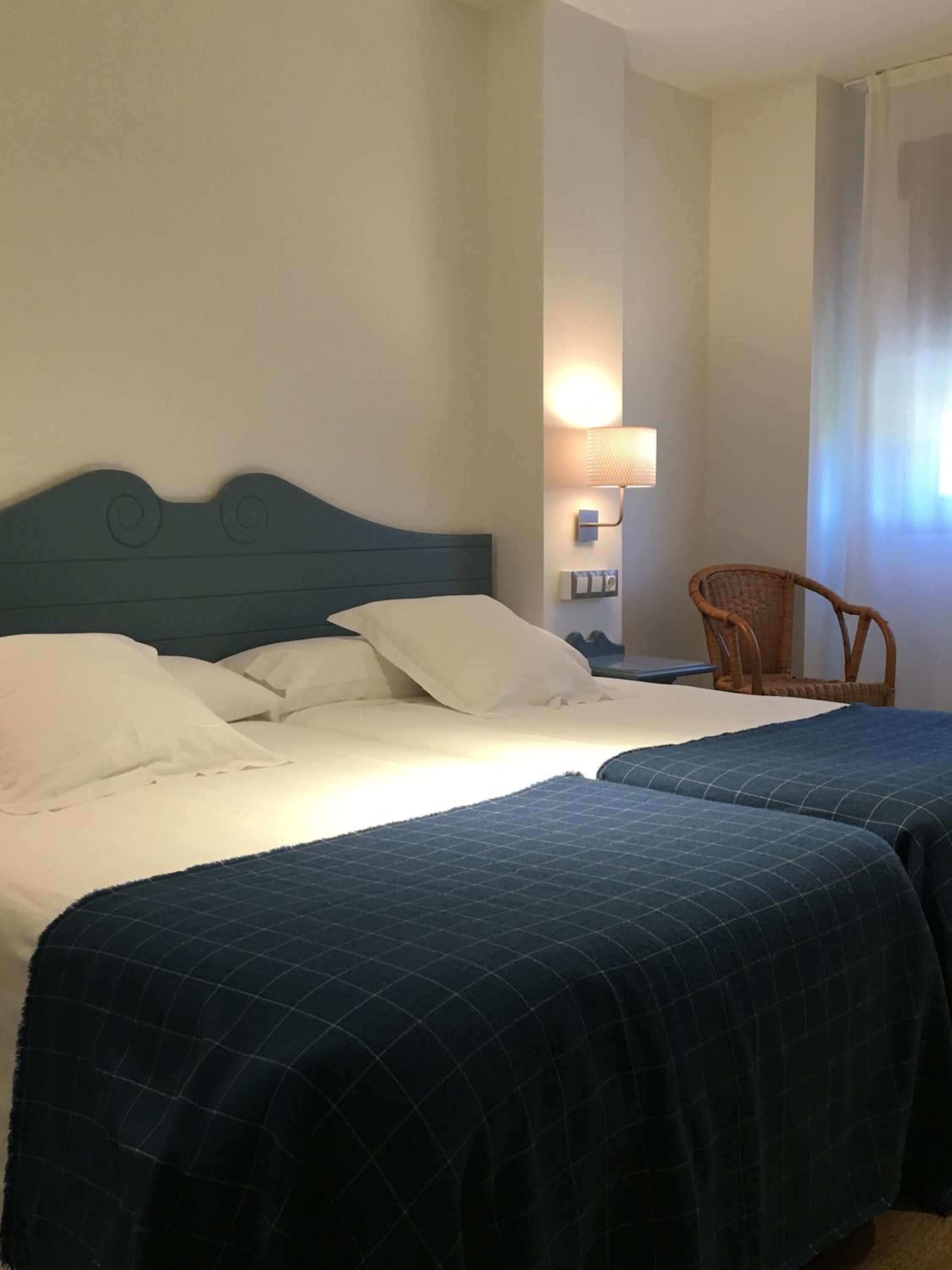 A hotel room with two beds, a nightstand, a wall-mounted lamp, a wicker chair, and a window with curtains.