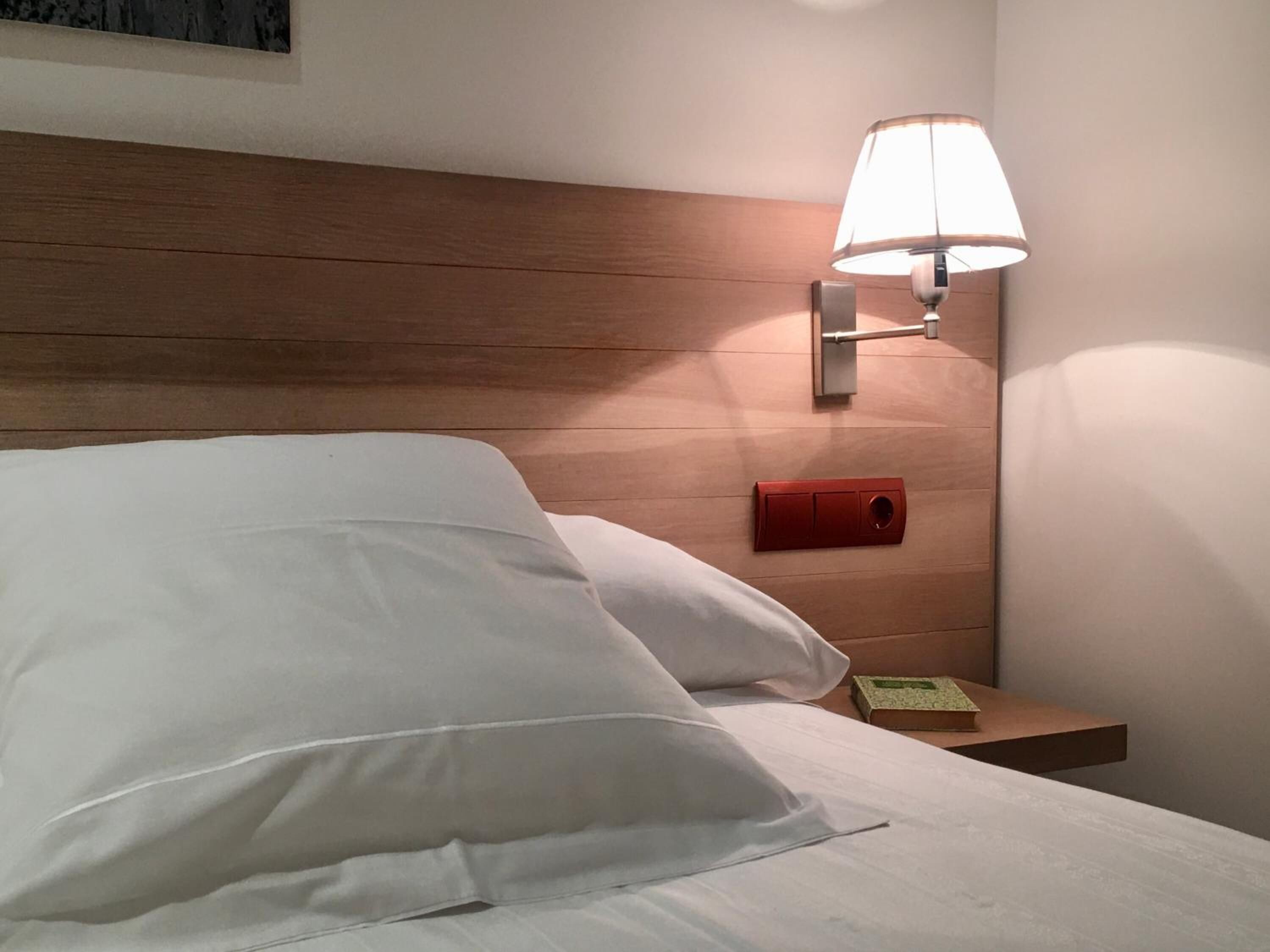 A hotel room with a wooden headboard, a bedside lamp, a nightstand with a red object, and a green book.