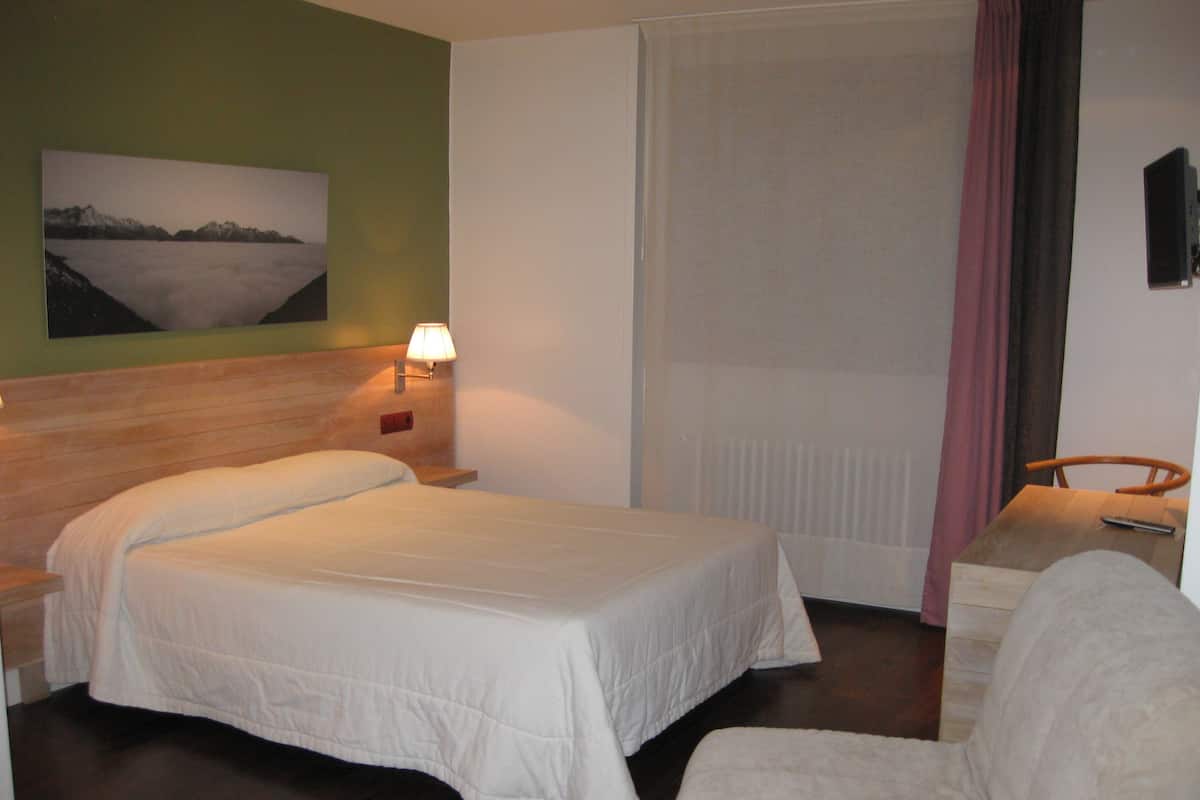 Quarto de hotel com cama, cadeira, mesa de cabeceira, abajur, quadro na parede e janela com cortinas.