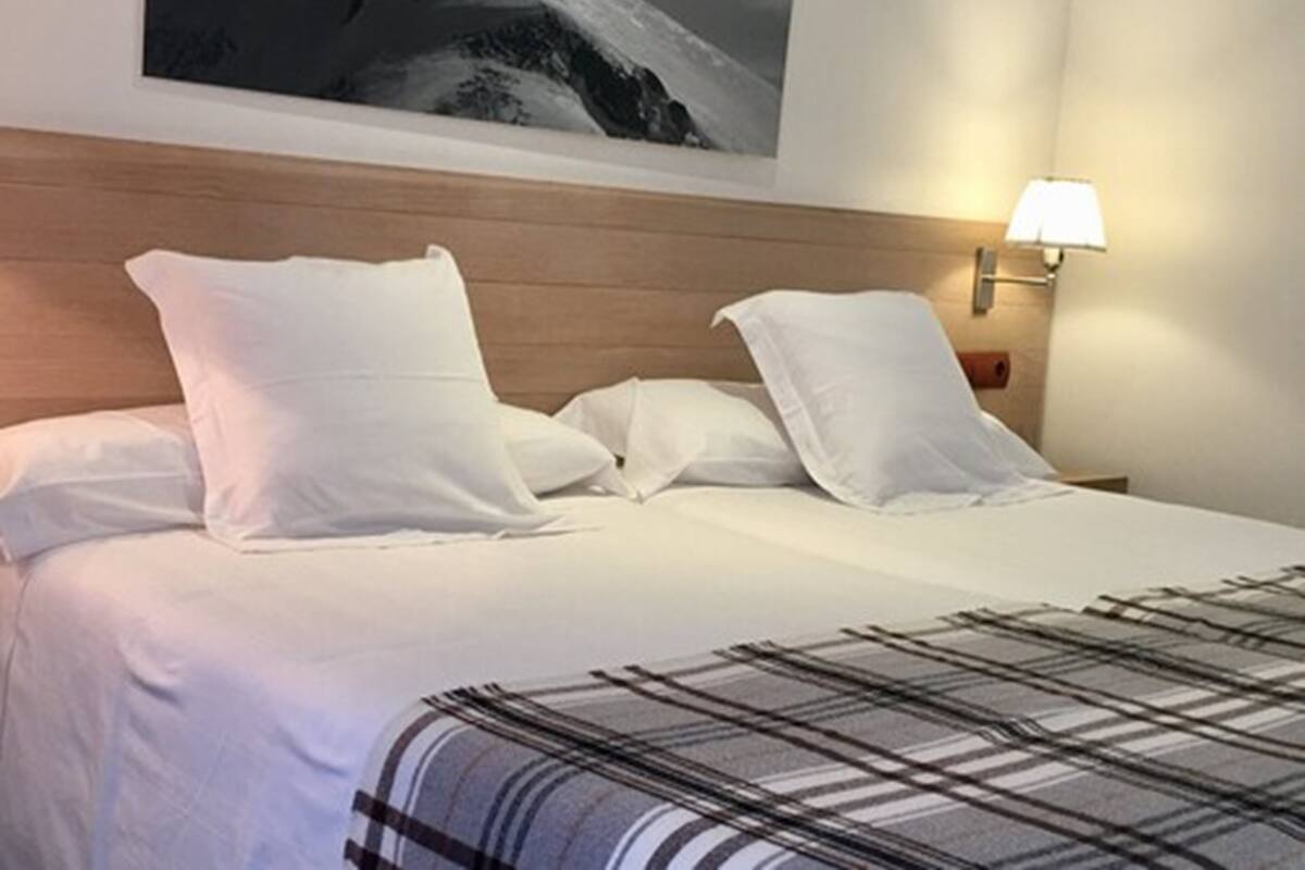 Cama bem arrumada com lençóis brancos e manta xadrez, cabeceira de madeira e luminária fixada na parede.