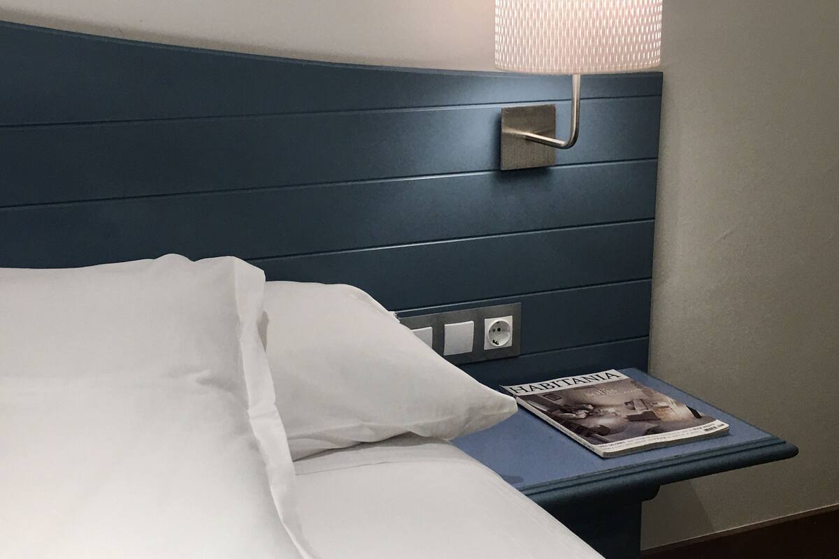 Quarto de hotel com uma cama, mesa de cabeceira, abajur e uma revista.