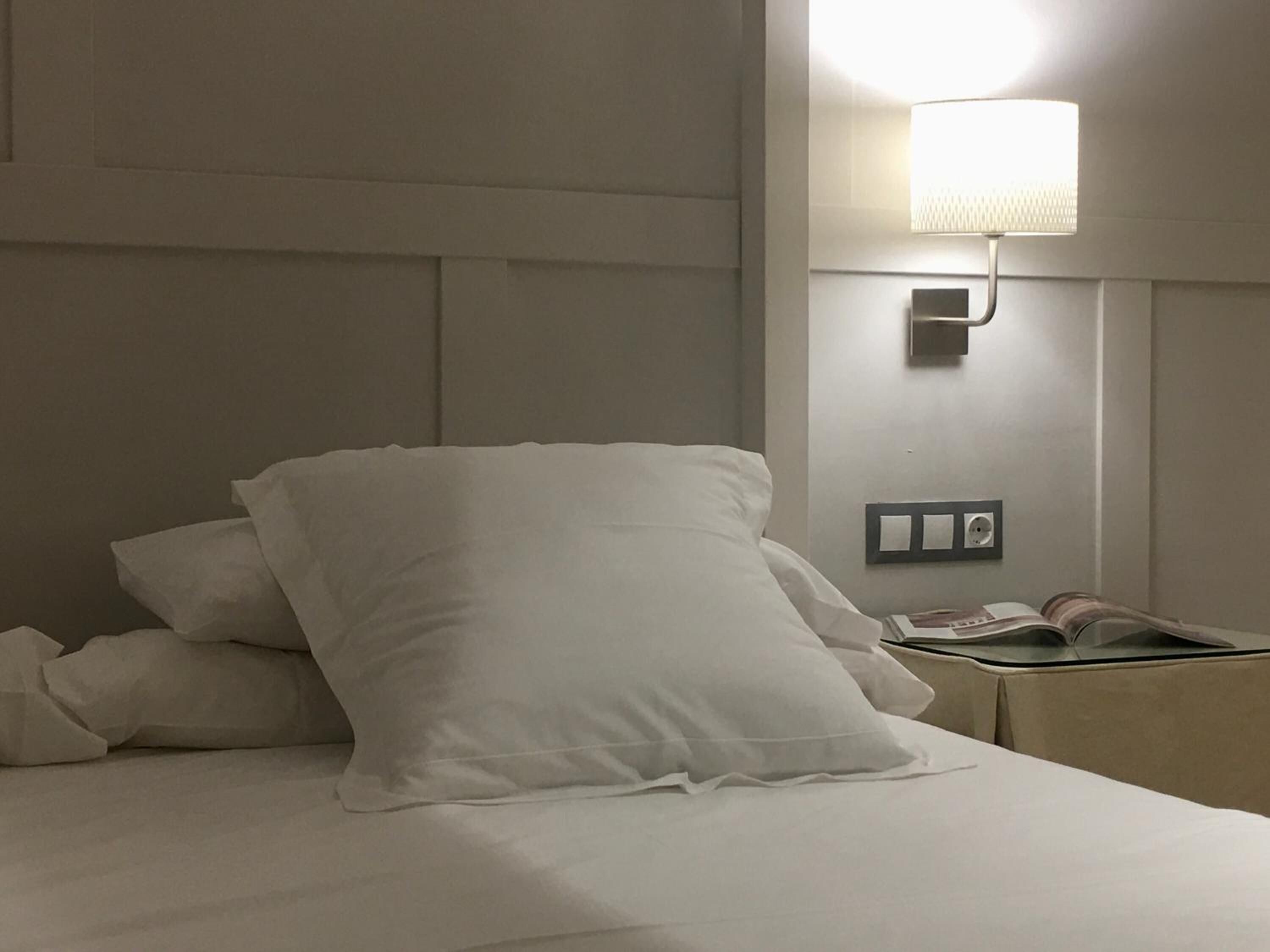 A neatly made bed with white bedding and pillows, a bedside table with a lamp, and a book.