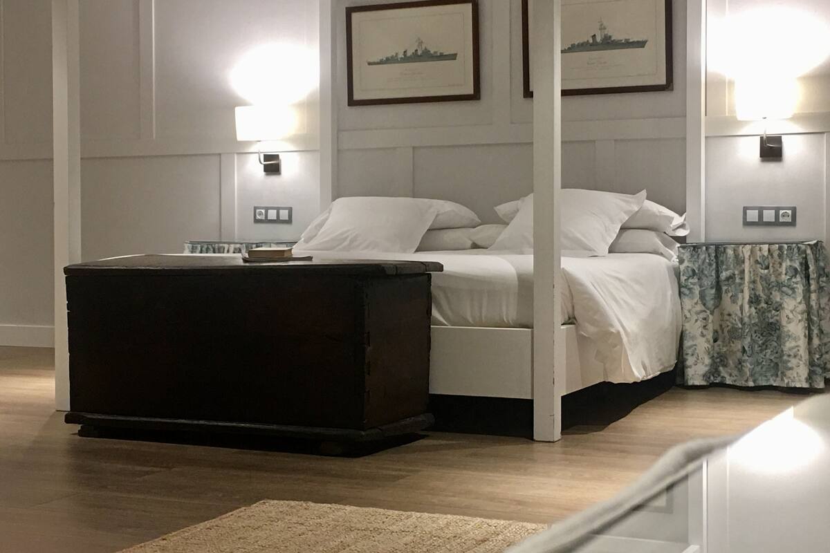 Quarto com cama, mesa de cabeceira, cômoda e duas luminárias fixadas na parede.