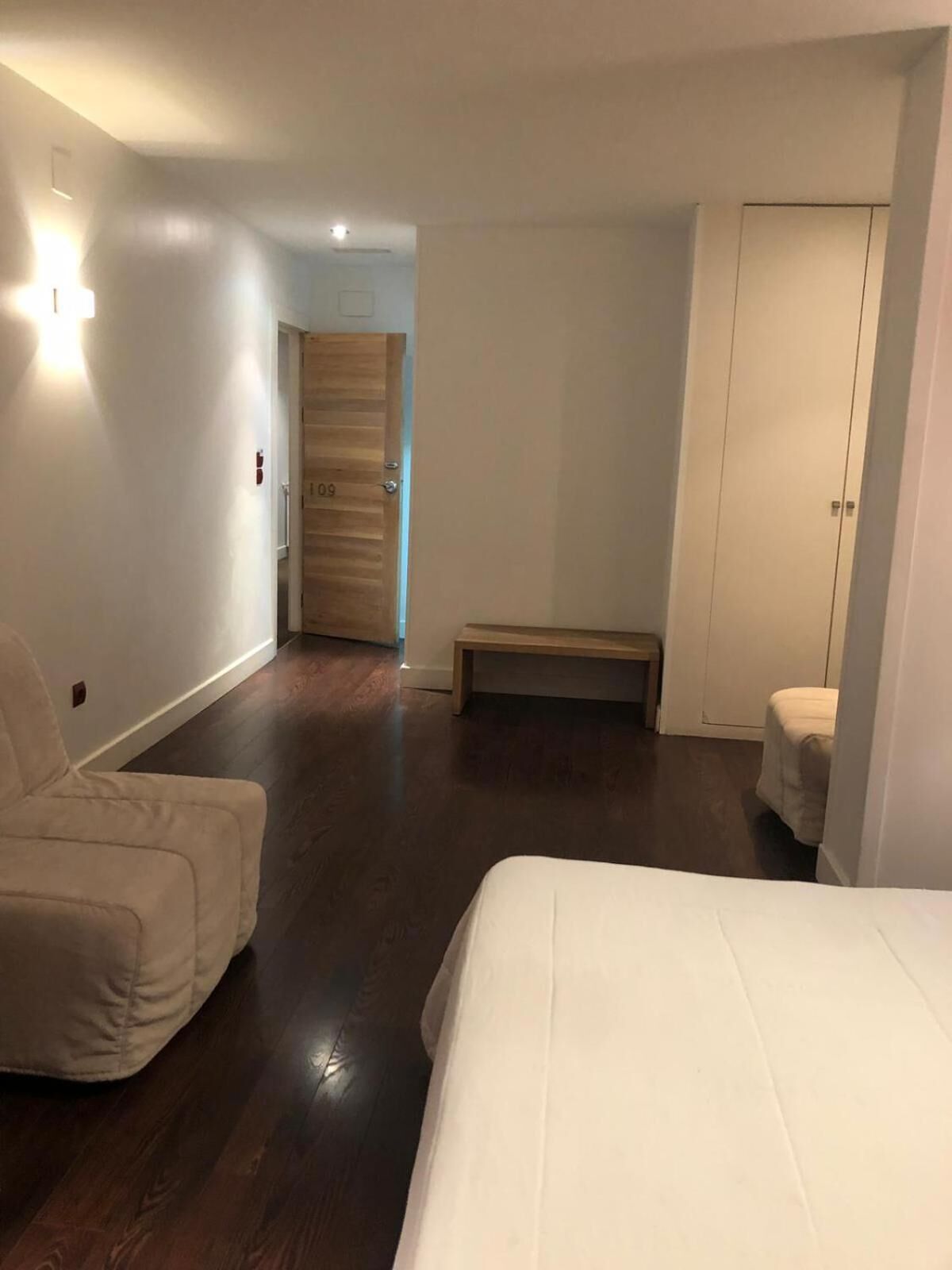 A hotel room with a bed, a chair, a bench, and a wardrobe.