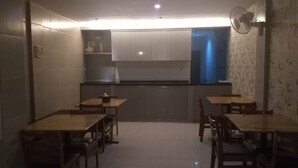 A dining area with wooden tables and chairs, a kitchen in the background, and a wall-mounted fan. - Permata Inn (Medan)
