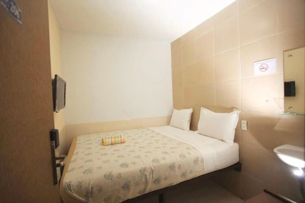 Una habitación pequeña con cama individual, una colcha floral color beige, una mesita de noche, un televisor montado en la pared y un baño con inodoro y lavabo.