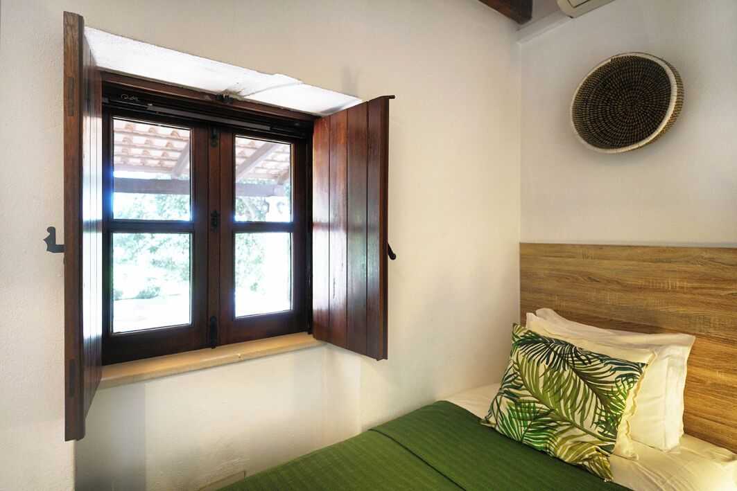 Una habitación con cama, ventana con contraventanas de madera y un respiradero en la pared.