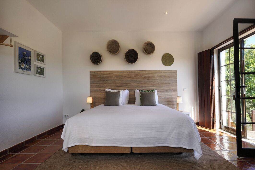 Une chambre avec un grand lit, une tête de lit en bois et une vue sur la verdure par la fenêtre.