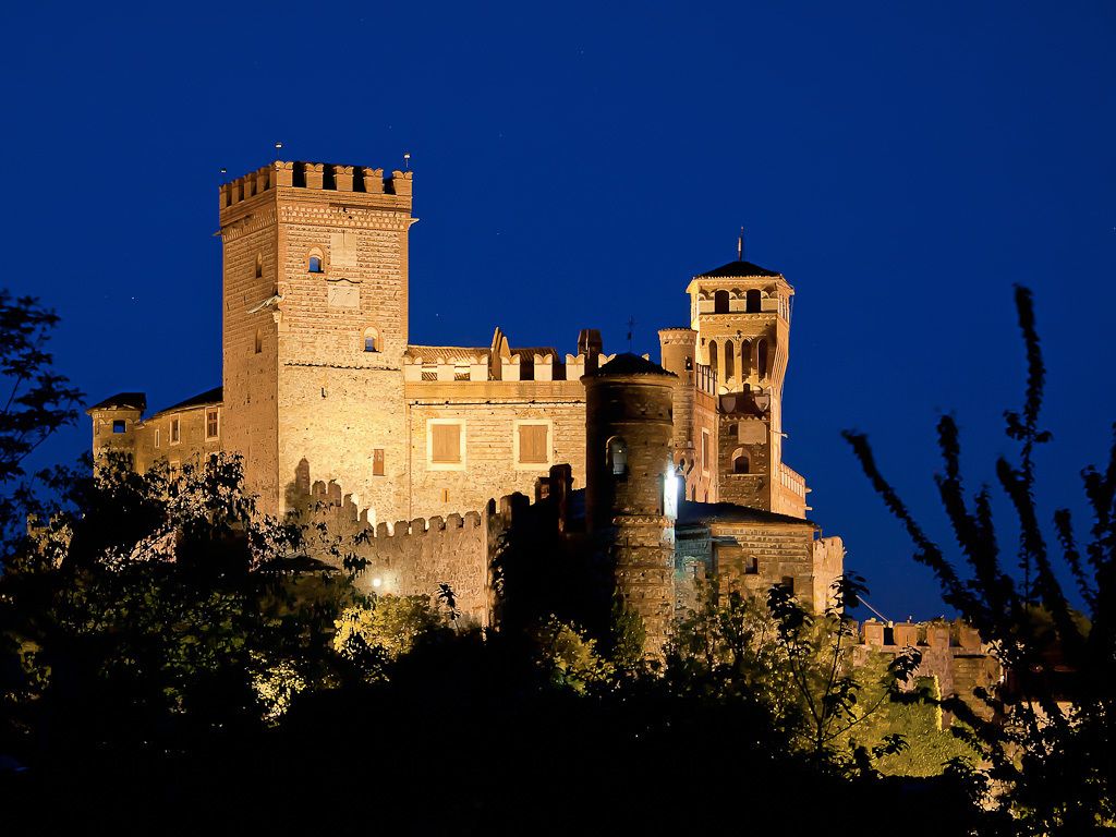 Un castillo histórico con una torre destacada y una torreón más pequeña, rodeado de árboles.