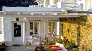 A white building with a stone wall, a covered patio with a table and chairs, and a view of the hills. - Vega Apartments (Tinos)