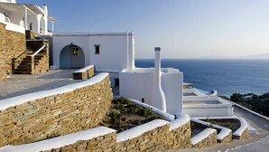 A white building with a stone wall and a view of the sea. - Vega Apartments (Tinos)