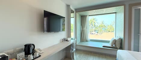 A hotel room with a bed, a TV mounted on the wall, a window with a view of palm trees, a desk with a coffee maker, and a chair.