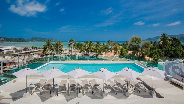 Área de piscina en la azotea con varios sillones de sol y sombrillas, con vistas a un paisaje costero con palmeras y un cielo azul despejado.