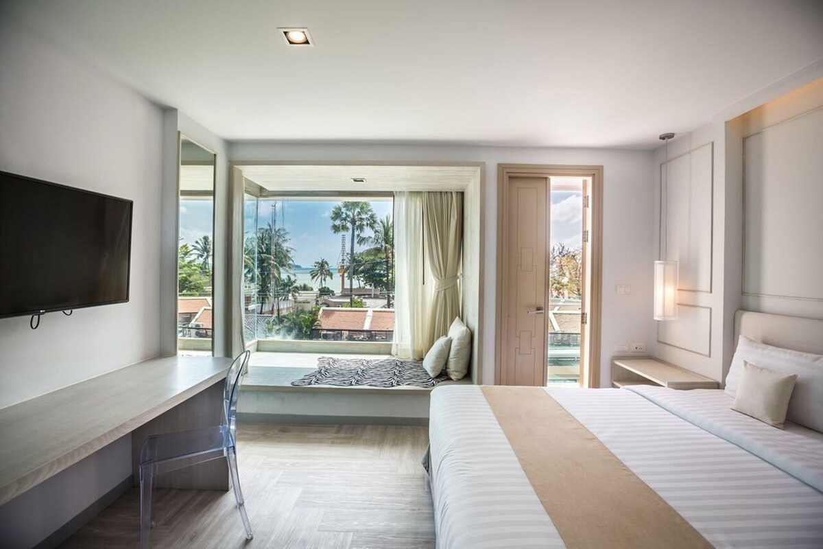 A hotel room with a bed, a desk, a TV, and a large window offering a view of palm trees and buildings.