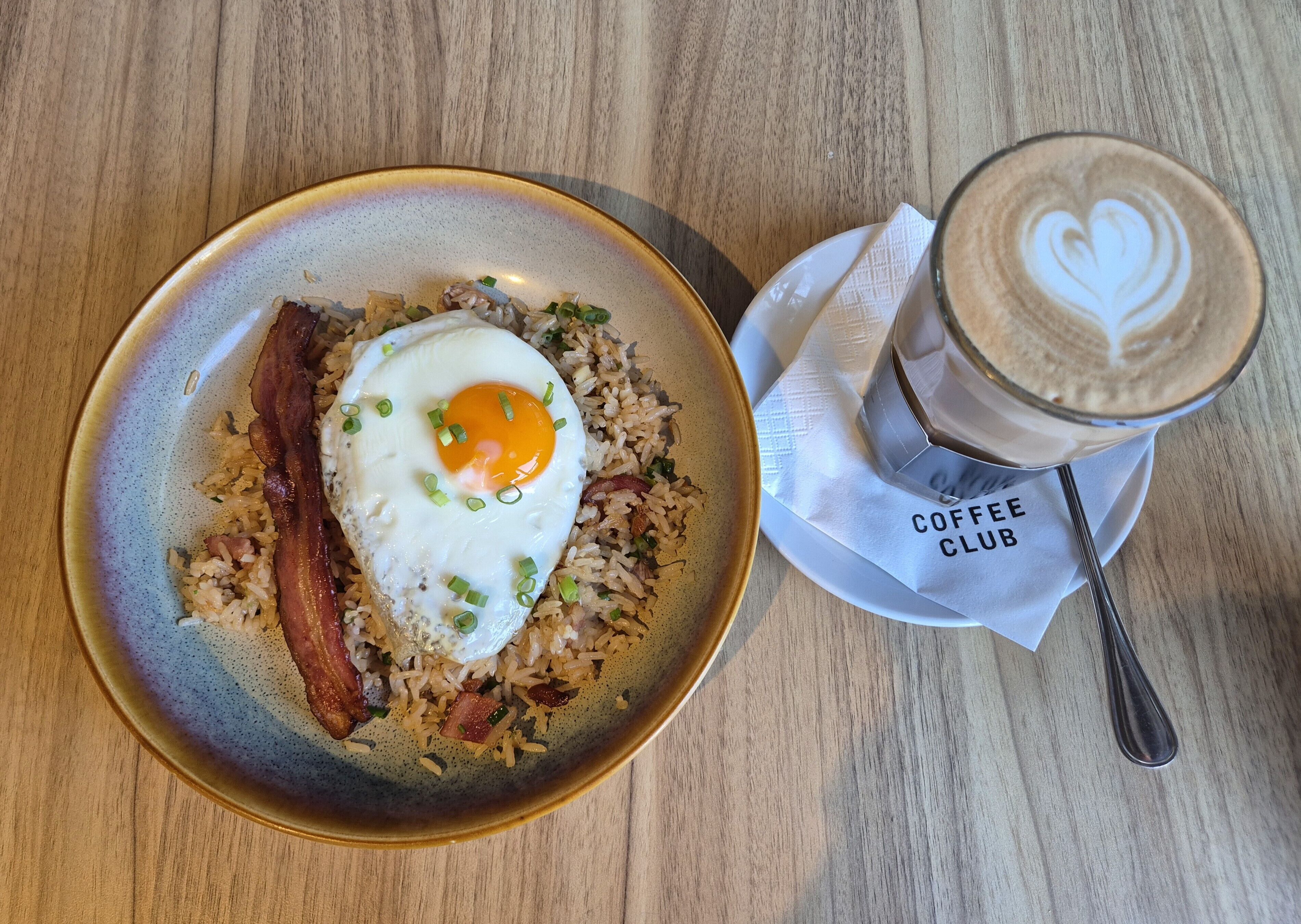 A plate of fried rice with a fried egg and bacon, and a cup of coffee with latte art on a wooden table.