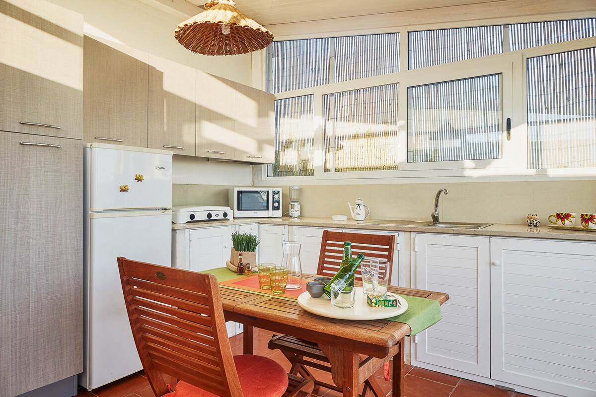 Cozinha compacta com área de refeições, equipada com geladeira, micro-ondas, pia e janela com persianas.