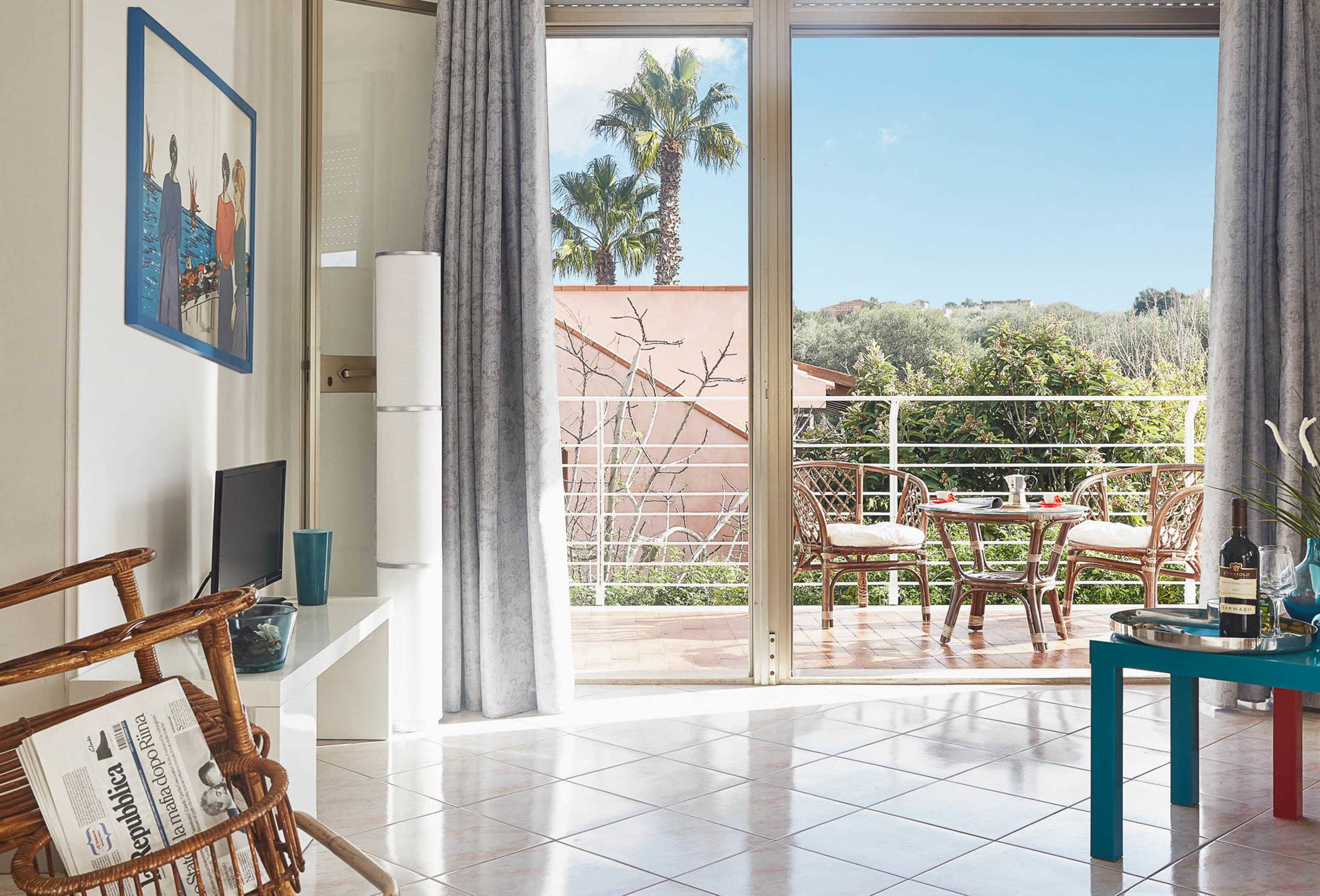 A modern living room with a balcony, wicker furniture, a dining area, and a view of palm trees and hills.