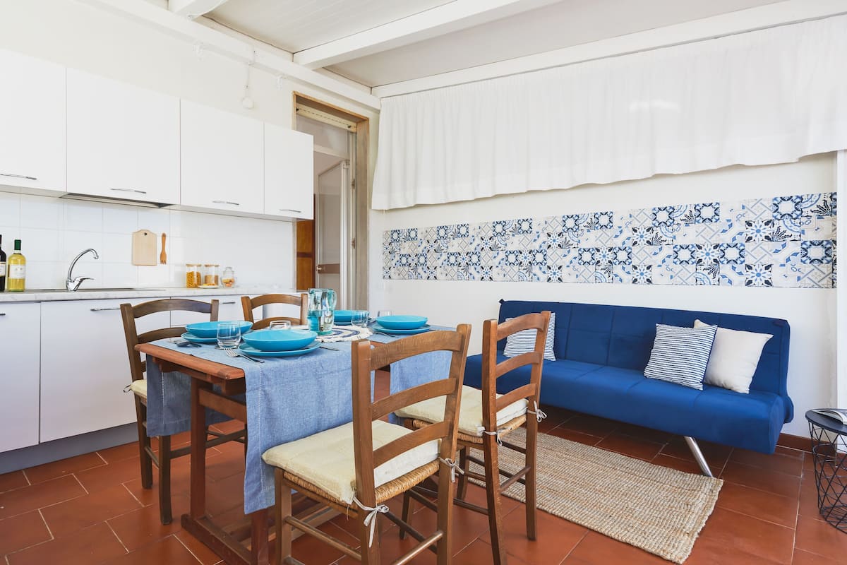 Uma cozinha compacta com armários brancos, uma mesa de jantar posta com pratos azuis e um sofá azul na sala de estar.