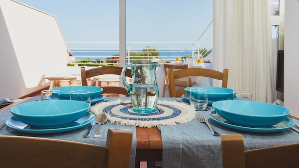 A dining table set with blue plates, a pitcher, and cutlery on a balcony overlooking the sea. - Villa Marina Green CaseSicule (Pozzallo)