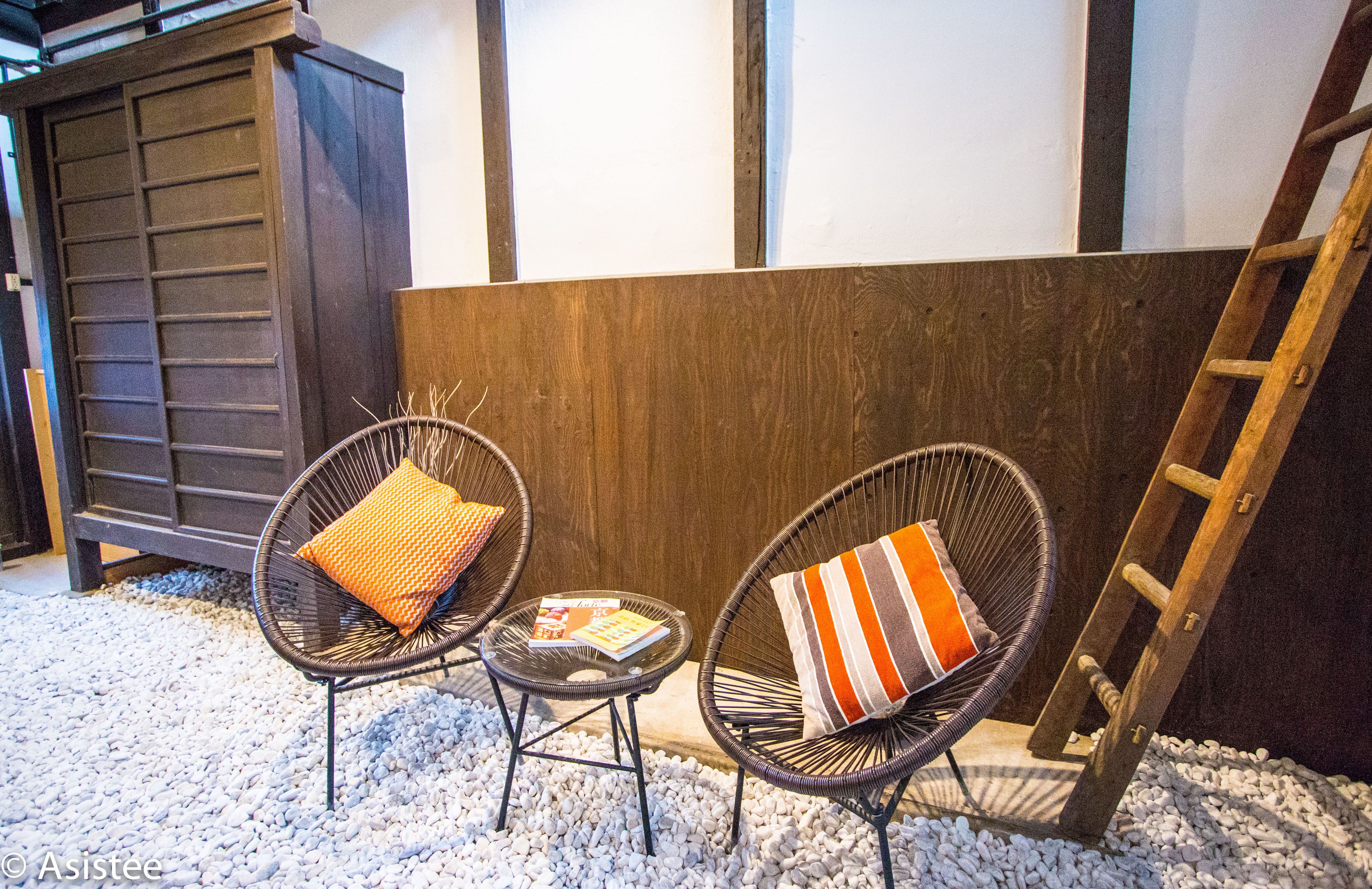 Deux fauteuils en osier avec des coussins, une petite table ronde avec un magazine, et une échelle en bois appuyée contre un mur orné d’un panneau en bois et d’une porte sombre.