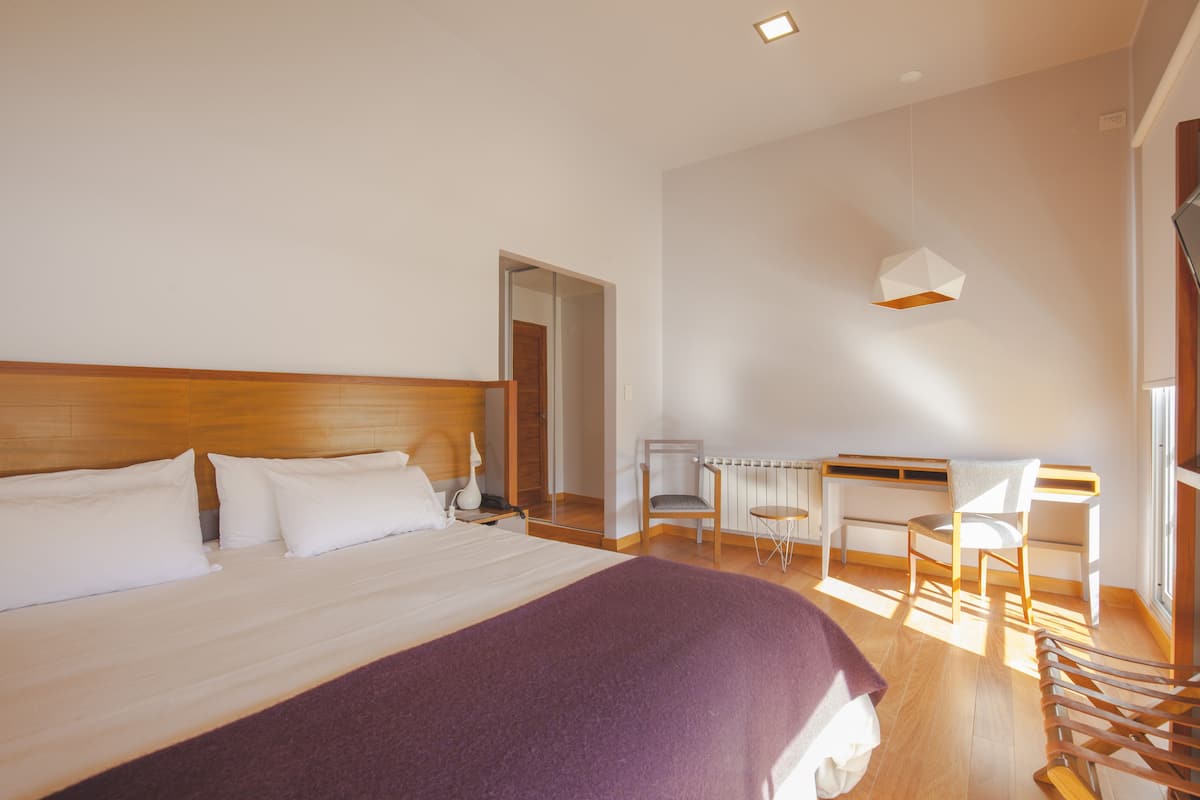 Une chambre d’hôtel moderne, dotée d’un grand lit, d’un petit bureau, de deux chaises et d’une fenêtre laissant entrer la lumière naturelle.