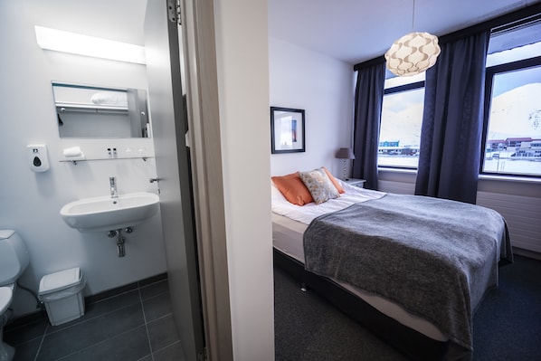 Habitación de hotel con baño y cama. El baño tiene lavamanos, inodoro y espejo. La cama tiene una manta gris y dos almohadas. Hay una ventana con vistas al exterior.