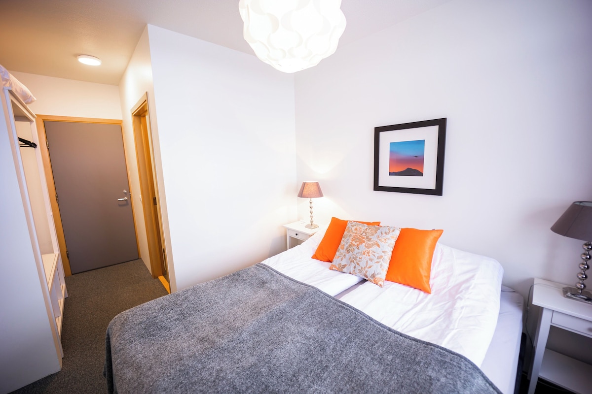 A neatly made bed with white and orange bedding, a grey throw blanket, and a framed picture on the wall.