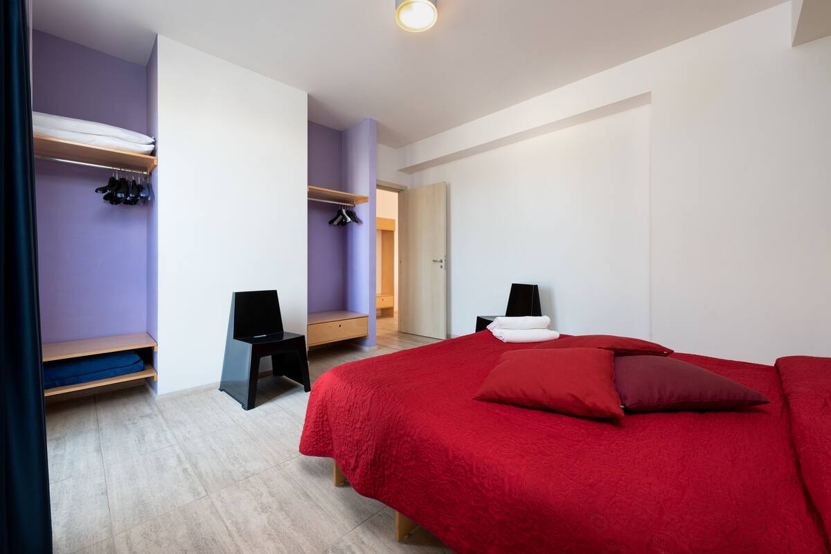 Ein Schlafzimmer mit einem roten Bett, einem Holzregal, einem Schreibtisch und einem Kleiderschrank.