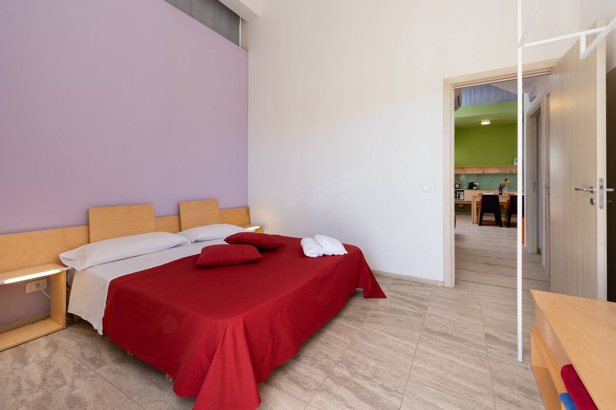 Ein ordentlich bezogenes Bett mit roter Bettwäsche und weißen Kissen, ein Nachttisch mit einer Lampe und eine Holzbank am Fußende des Bettes.